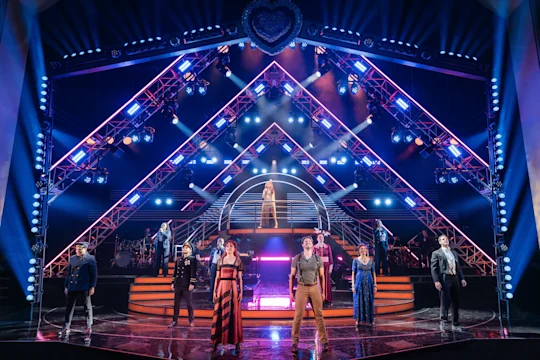 A group of performers stands on a brightly lit stage with a geometric, illuminated set and a singer elevated at the center.