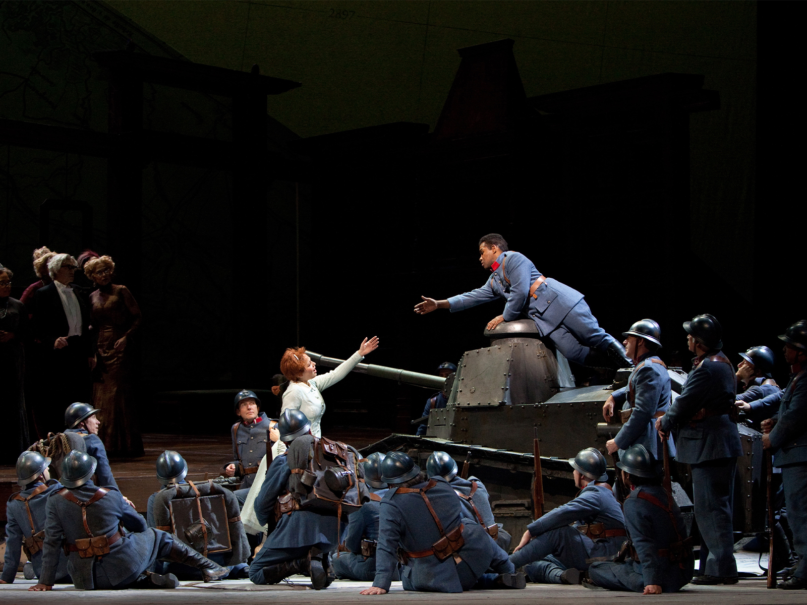 A group of soldiers in blue uniforms surrounds a tank on stage, with a woman in white reaching toward a man on top of the tank during a theatrical performance.