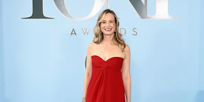 A woman in a strapless red dress stands smiling in front of a blue backdrop with "TONY AWARDS" written on it.