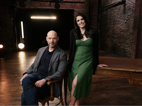 A man sits on a chair next to a wooden table while a woman in a green dress stands beside him in a dimly lit room with brick walls and wooden floors.