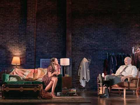 A woman sits on a green couch talking to a man in a chair on a stage set with brick walls, lamps, clothes rack, and a trunk, suggesting a cozy living room scene.