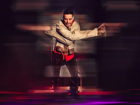 A person in a white jacket and red shirt performs a dynamic dance move with blurred motion effects, arms crossed in front of them.