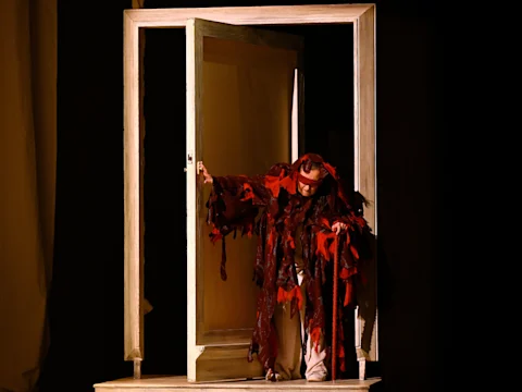A performer in a tattered, red and brown costume stands hunched over with a cane, emerging from a doorframe on a dimly lit stage.
