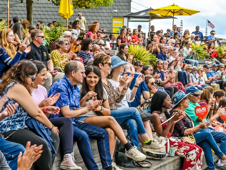 A large group of people sit on outdoor bleachers, clapping and watching an event on a sunny day, with greenery and umbrellas in the background.