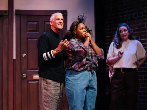 Three people are on stage performing. An elderly man gestures while beside a young woman who looks apprehensive, and another woman stands behind them watching.