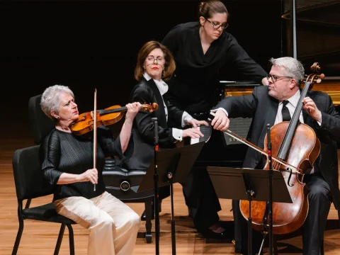 Four musicians perform on stage; an older woman plays the violin, a younger woman assists with sheet music at a piano, and a man plays the cello while a seated woman watches intently.