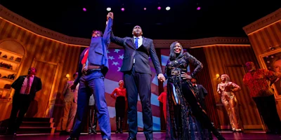 A group of people on a stage. Two men and a woman stand in front in formal attire; the men hold each other's hands up in the air while the woman strikes a pose. An American flag is projected in the background.