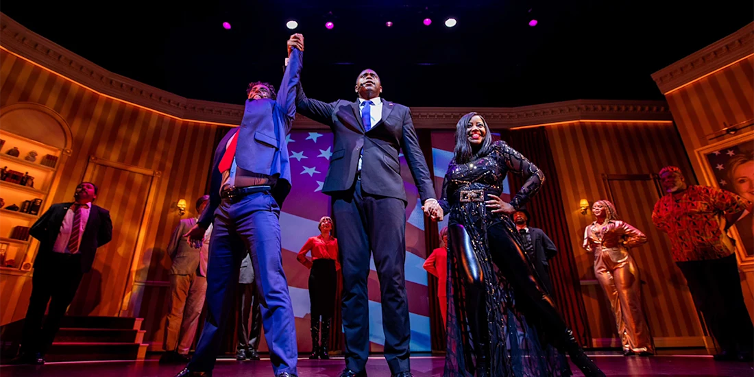 A group of people on a stage. Two men and a woman stand in front in formal attire; the men hold each other's hands up in the air while the woman strikes a pose. An American flag is projected in the background.