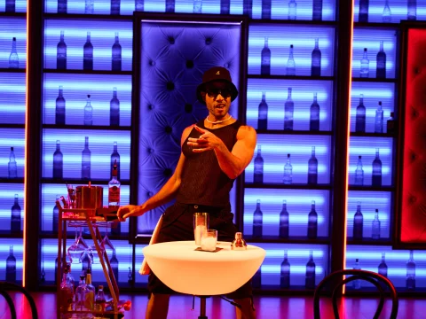 A person wearing a hat and sunglasses gestures behind a small table in front of a colorful wall with bottle displays.