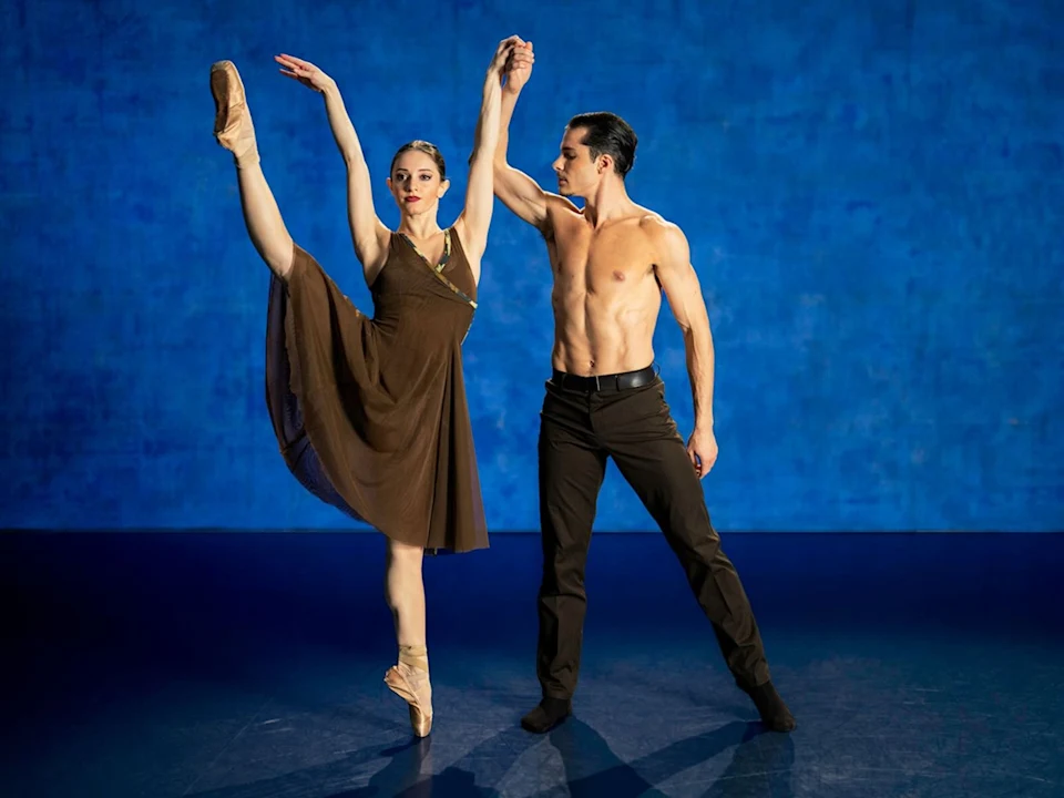 A female ballet dancer performs an arabesque while a male dancer stands beside her, holding her hand, against a blue background.