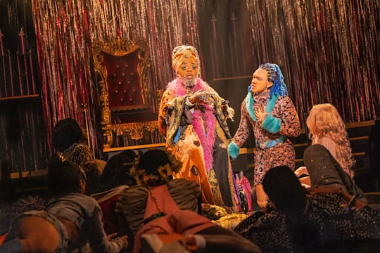 Actors in colorful costumes perform on stage with a throne and metallic streamers in the background, surrounded by people seated or kneeling in the foreground.
