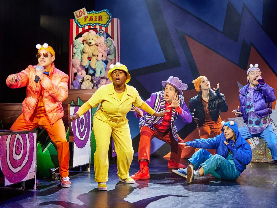 A vibrant stage performance with six actors in colorful costumes animatedly gesturing and posing.