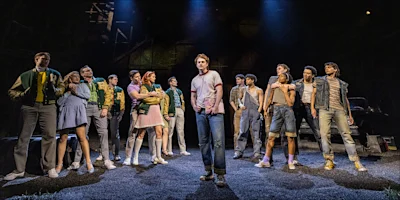 A group of actors in casual and vintage clothing stands on stage under dramatic lighting, facing forward, as part of a theater production.