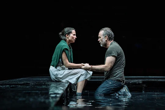 A woman sits on a platform while a man kneels in water, both holding hands and facing each other in a dramatic, dimly lit setting.