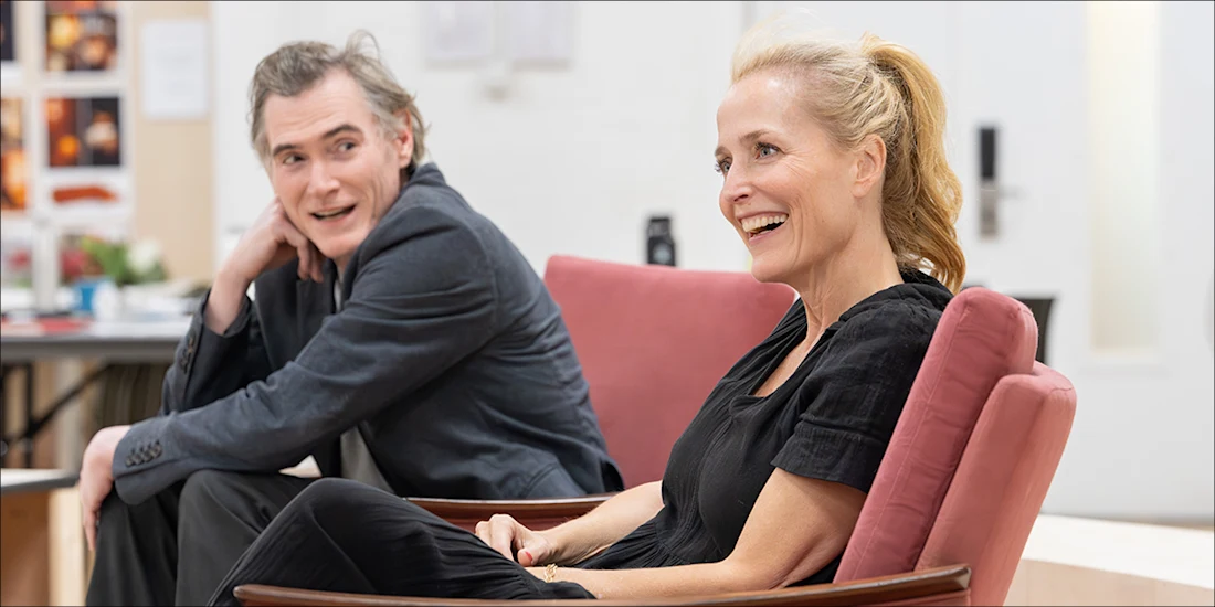 Two adults sit on red chairs in a bright room, engaged in conversation and smiling. Office materials and wall displays are visible in the background.