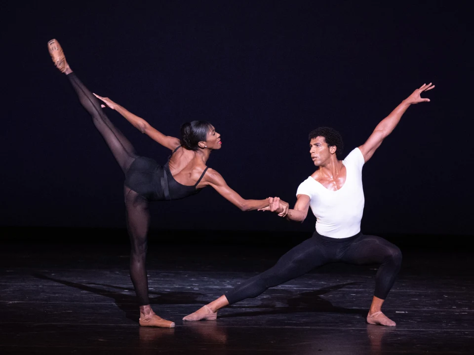 Production photo of Pathways to Performance in Washington, showing two ballet dancers perform on stage, one extending a leg high while the other poses in a low lunge, both wearing black tights.