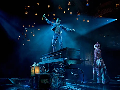A man stands on a wooden cart holding an object aloft, illuminated by blue stage lights, while a woman in period costume looks on; lanterns hang above the scene.