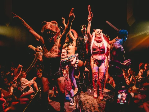 A group of individuals in elaborate costumes, including a dinosaur headpiece and fantasy attire, perform energetically on a stage with an audience clapping and cheering in the background.