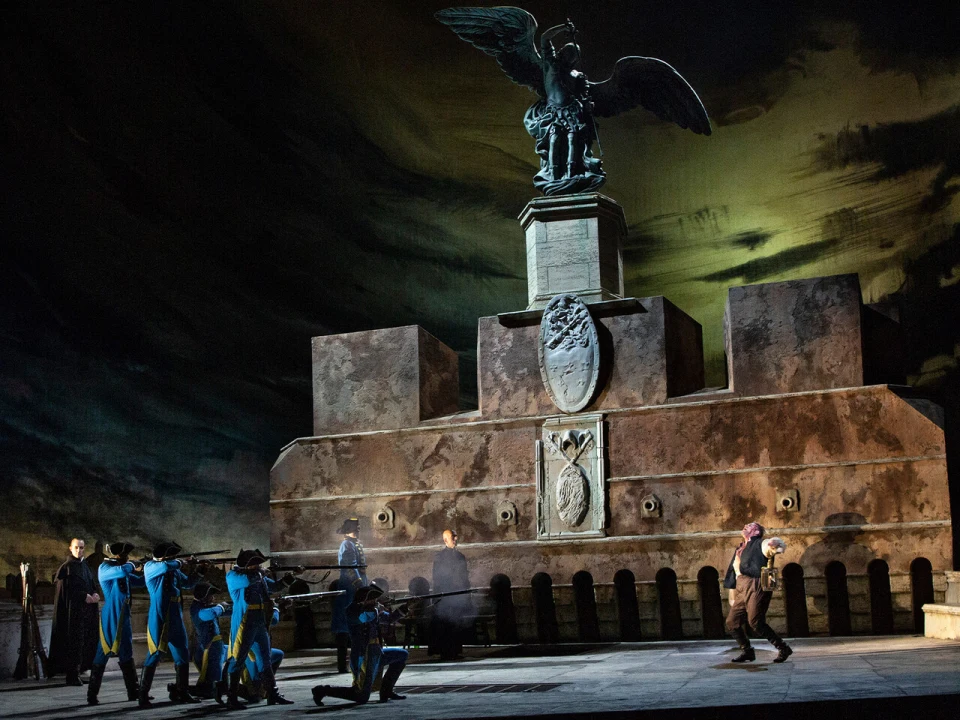 Production photo of Tosca, showing a dramatic stage performance with actors in period costumes beside a large, winged statue backdrop featuring dark, intense lighting.