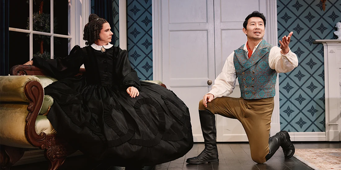 A woman in a black Victorian dress sits on a sofa while a man in period costume kneels on one knee, gesturing with his right hand in a dramatic stage setting.