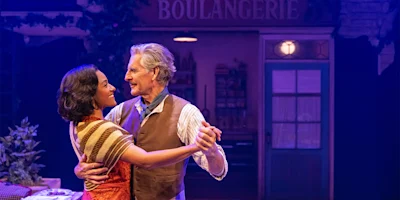 An older man and a younger woman are dancing together and smiling on a stage set resembling a bakery, with warm lighting and a sign reading "BOULANGERIE" in the background.