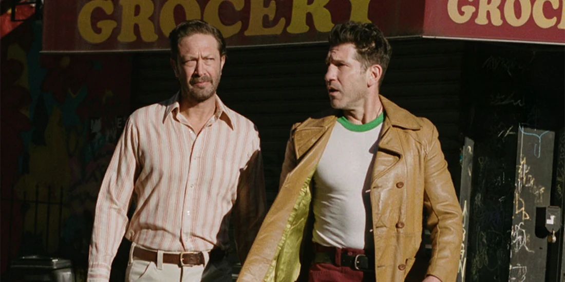 Two men walk side by side on a street in front of a grocery store; one wears a striped shirt, the other a yellow jacket over a white shirt.