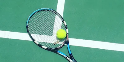 A tennis racket and a yellow tennis ball rest on a green court near intersecting white boundary lines.