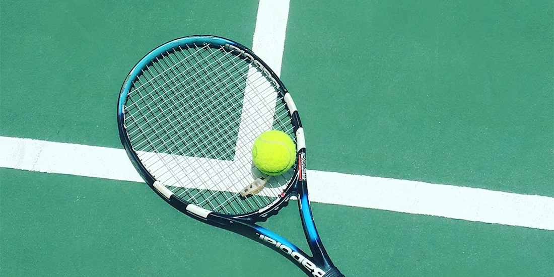 A tennis racket and a yellow tennis ball rest on a green court near intersecting white boundary lines.