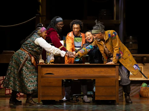Five actors on stage stand around a wooden desk, reaching their hands together in the center as if in a group gesture, with theatrical props and costumes visible.