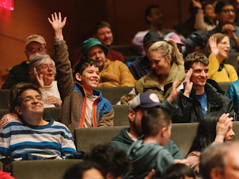 A group of people sit in an auditorium, with several individuals raising their hands; one boy in the center smiles with his hand raised.