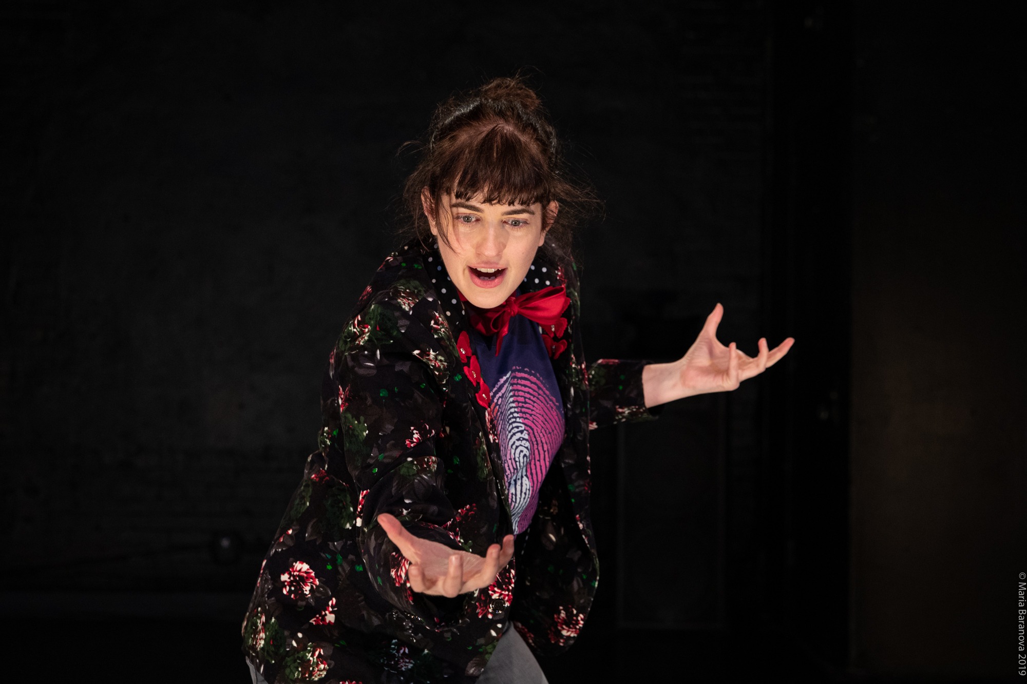 A person with brown hair and bangs, wearing a floral jacket and red scarf, gestures expressively with both hands in front of a dark background.