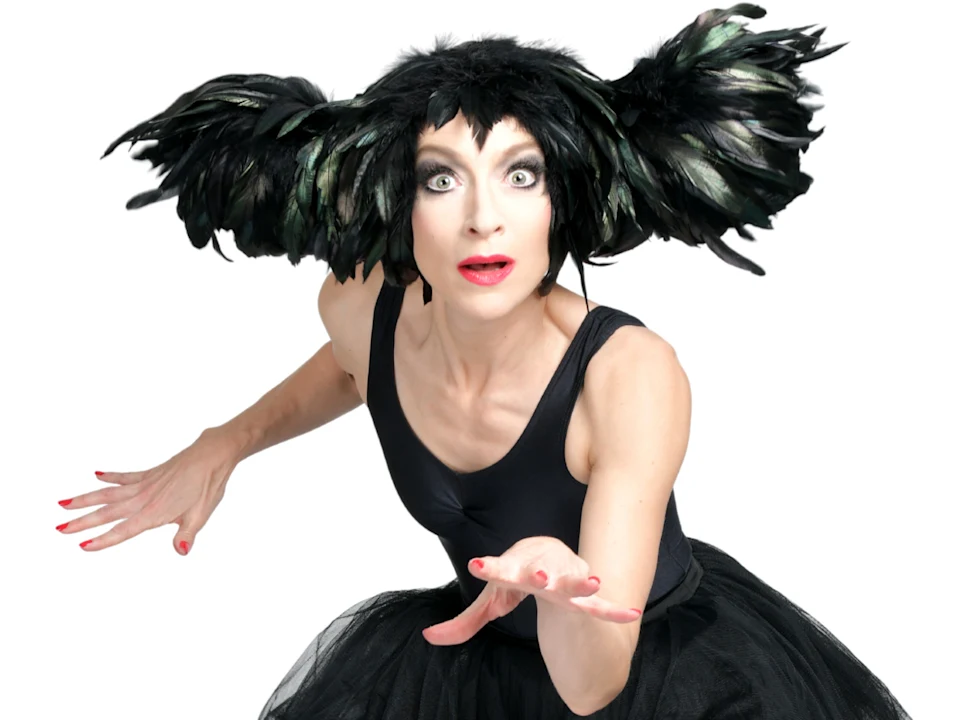 A person in a black leotard and tutu wearing a large, wing-like feathered headpiece, looking surprised with hands outstretched.