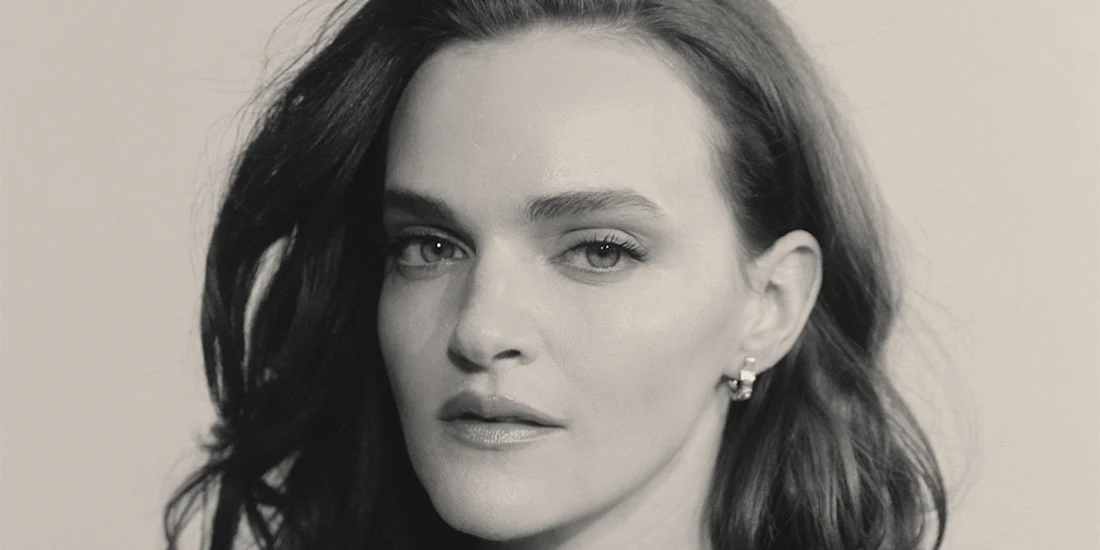 A woman with long, wavy dark hair looks directly at the camera with a neutral expression. She is wearing small hoop earrings. The image is in black and white.