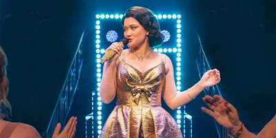 A woman in a gold dress sings into a microphone on stage, with bright lights behind her and audience members in the foreground.
