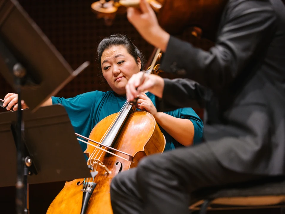 A cellist in a teal top performs with a violinist in a black suit during a musical concert, both focused on their instruments.