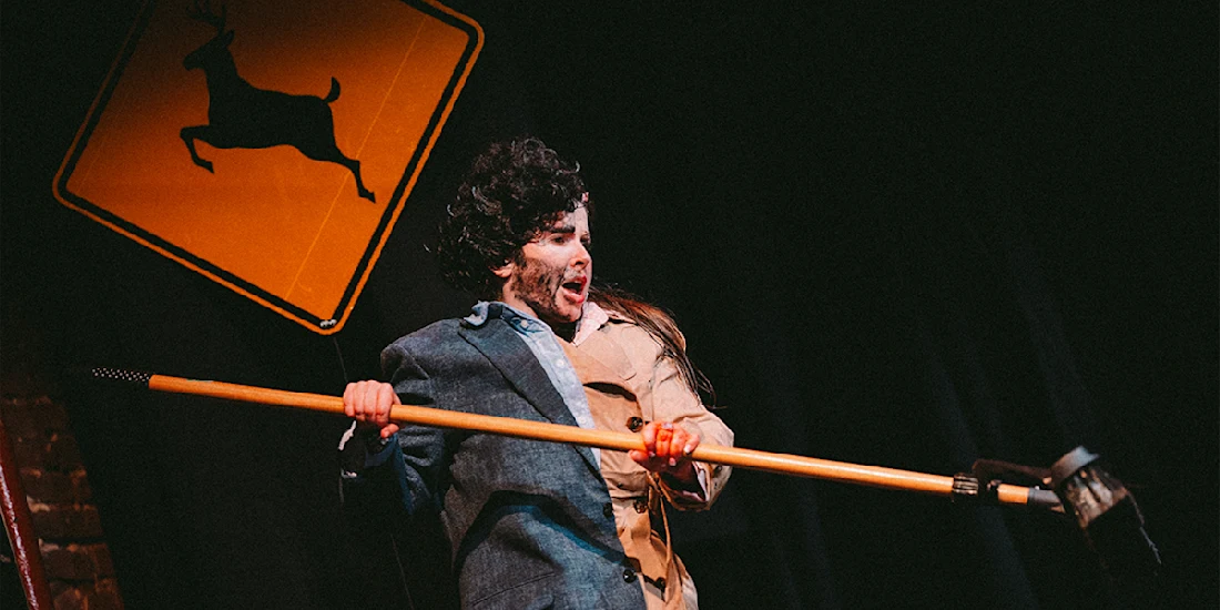 A person in a messy wig and costume holds a broom on stage, with a deer crossing sign in the background.