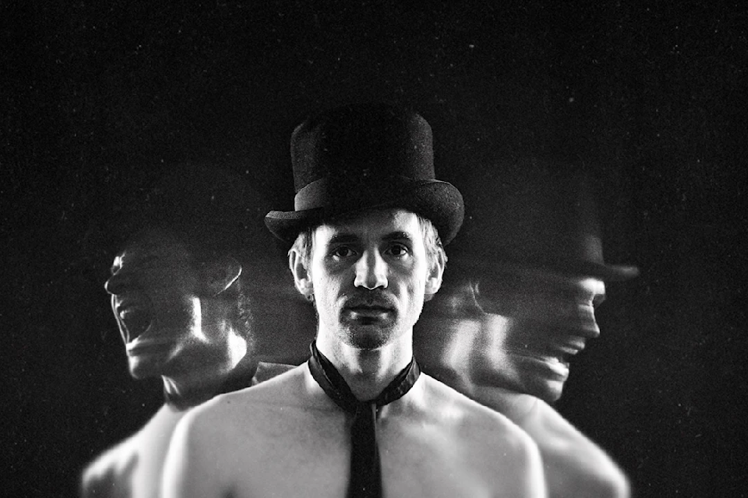 A shirtless person wearing a top hat and loose necktie stands against a dark background, with blurred duplicate images of their head on either side, suggesting motion or emotion.