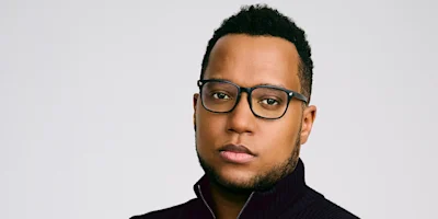 A man with short curly hair, wearing black rectangular glasses and a dark zip-up sweater, looks directly at the camera with a neutral expression against a plain background.