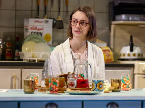 A woman wearing glasses and a white shirt sits at a blue table in a kitchen. The table has colorful dishes and glasses. The kitchen counter in the background has various items, including a cereal box and a dish rack.