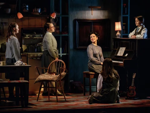 A group of five women in a dimly lit room, one at a piano, others standing or sitting, engaged in conversation. The setting is domestic with kitchen items visible.