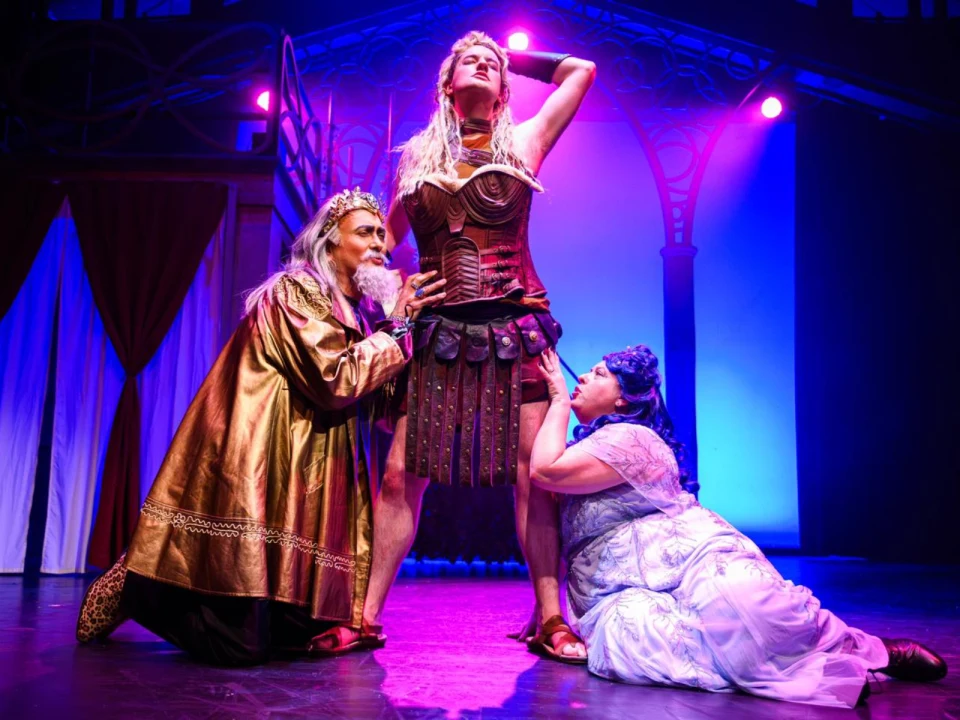 Three actors on stage in a dramatic scene: one dressed as a warrior standing confidently, two others kneeling and reaching toward the warrior. The background is illuminated with colorful stage lights.