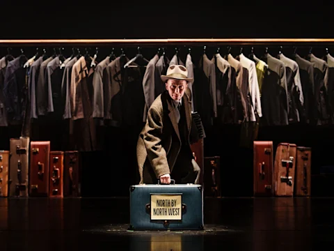A person in a suit and hat kneels, holding a suitcase labeled “North by Northwest,” with suit jackets and luggage in the background on stage.