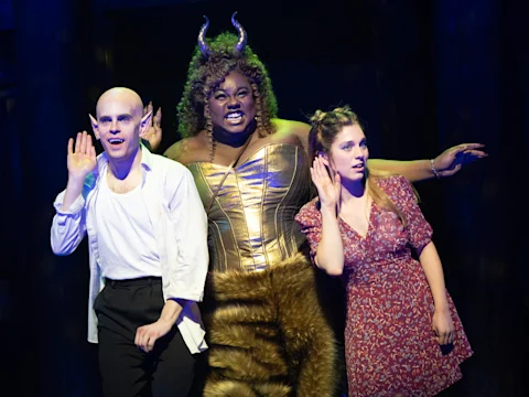 Three actors on stage in costume, two with elf ears and one dressed as a faun with horns, all holding a hand to their ear as if listening intently.
