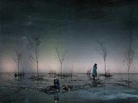 A large group of people dressed in period costumes dance in pairs in a grand, dimly lit ballroom with a chandelier and high ceilings.