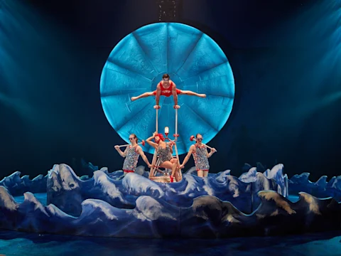 Performers balance on a pyramid structure with a circular blue backdrop, surrounded by blue wave-like decorations on stage.