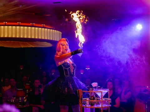 A performer in a black costume holds up a flaming object on stage, with an audience seated in the background under colorful lighting.