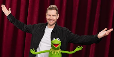 A man in a black jacket and white shirt stands in front of red curtains with arms outstretched next to Kermit the Frog, who is also posing with arms open.