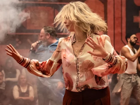 A woman with blonde hair dances energetically in a floral blouse. People are seated in the background, and a man holds a microphone amid a smoky atmosphere.