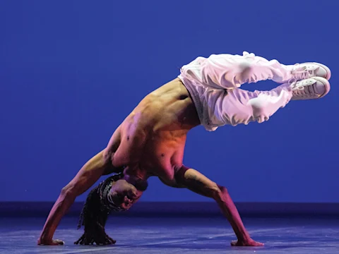 A dancer performs an inverted pose on stage, balancing on one hand with legs raised, wearing white pants and sneakers against a blue background.