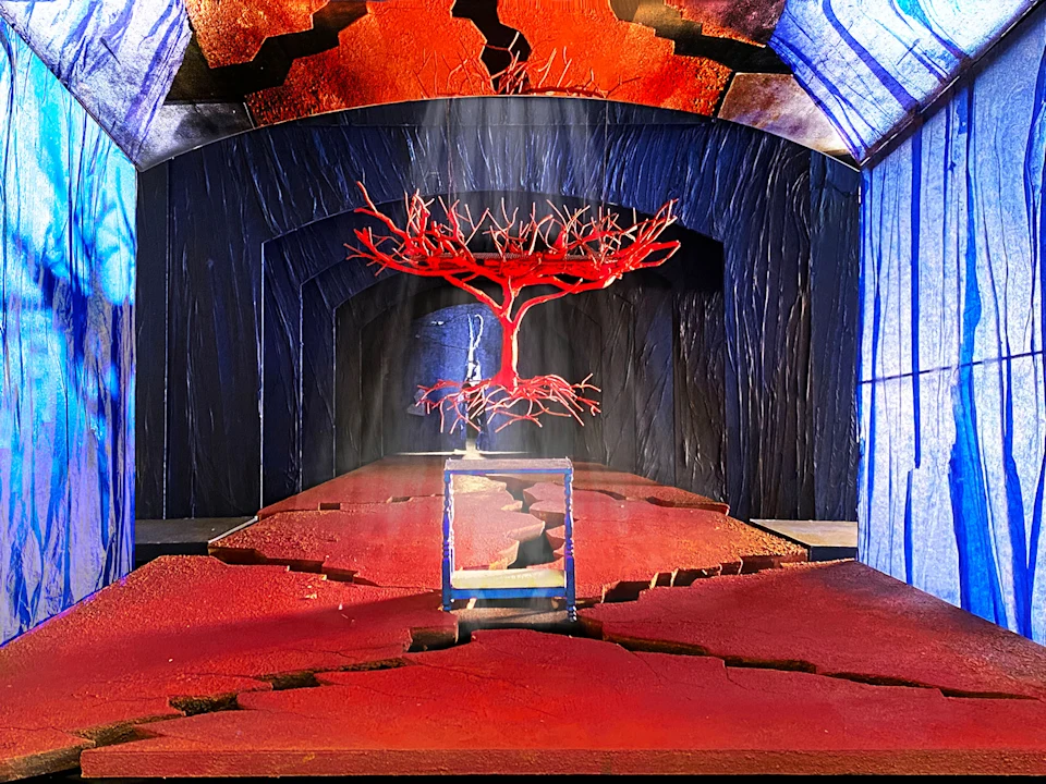 Production photo of El Último Sueño de Frida y Diego in New York, showing a red, leafless tree with exposed roots floats above a cracked red pathway in a tunnel with blue walls and ceiling featuring fissures.